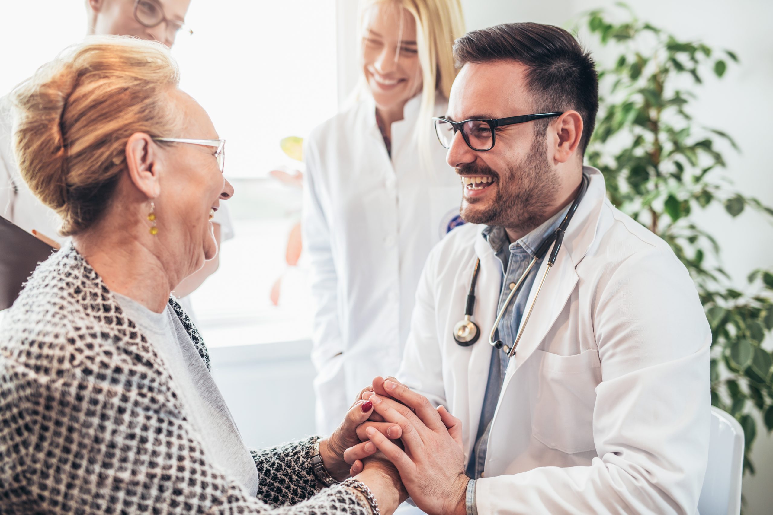 A doctor, embodying stress-free healthcare practices, smiles while holding hands with an elderly woman in a medical office.